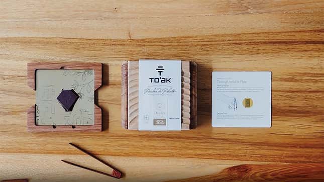 Three items laid out on a wood table: a metalic-plated tasting platter with a piece of chocolate on it, a wooden box, and a paper with writing on it
